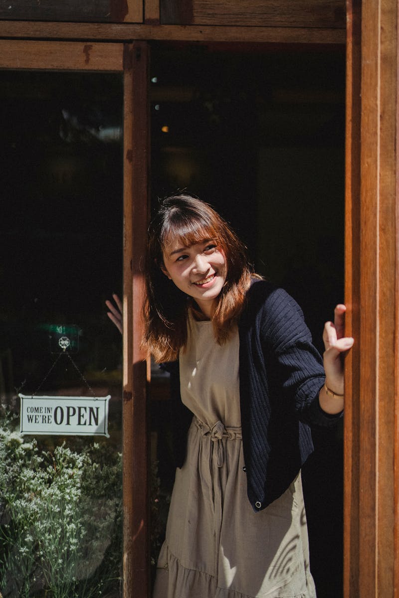 Positive smiling Asian female store worker opening store door and looking away contentedly